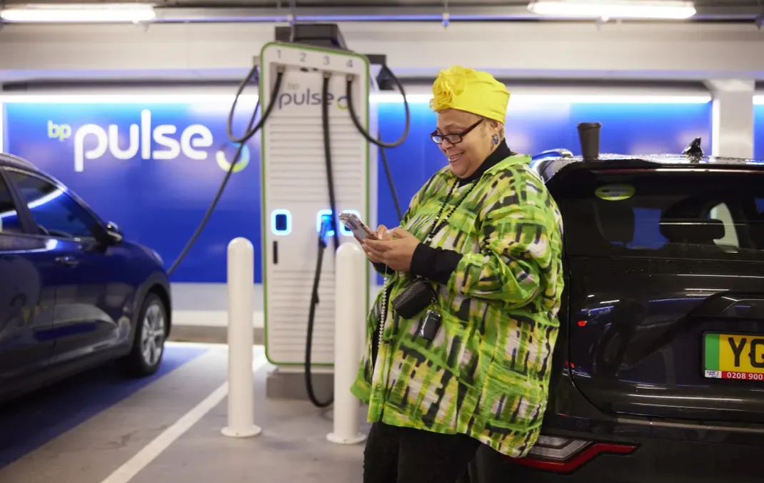 A woman is looking at her phone and smiling while her car is being charged at a bp pulse charging station.
