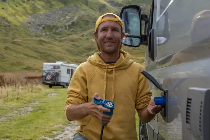 A photo of a man plugging his camper van in to charge while on a road trip in the mountains.
