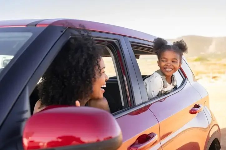 A young woman and her daughter are in a car on a road trip. The woman is looking back at her daughter who is smiling with her head out of the car.