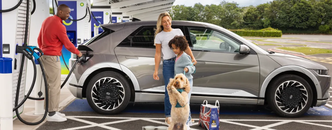 Two parents and their child, and poodle-mix dog, stand by their EV, as the dad is charging the car. 