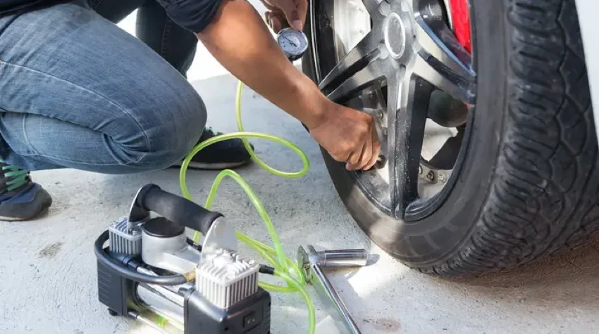 A man is kneeling down next to a vehicle wheel, inflating the wheel and he is holding a pressure gauge in his hand.