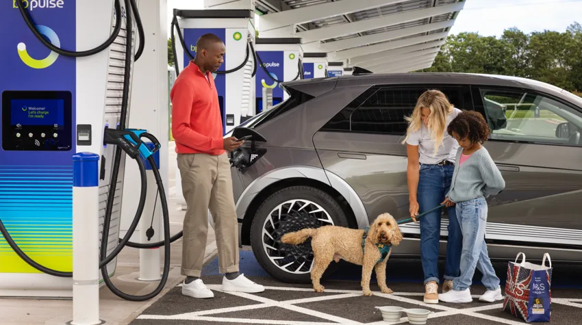 A man, woman, child and a dog stand by a silver EV as it charges. 