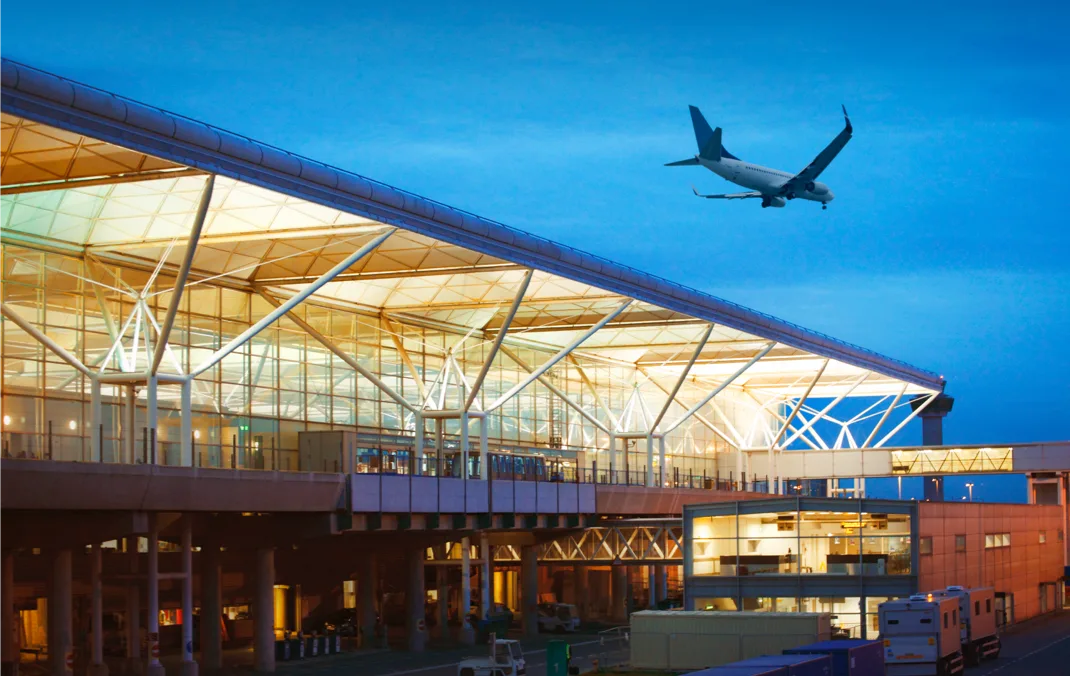 A plain is flying above the well lit Stansted airport terminal building.