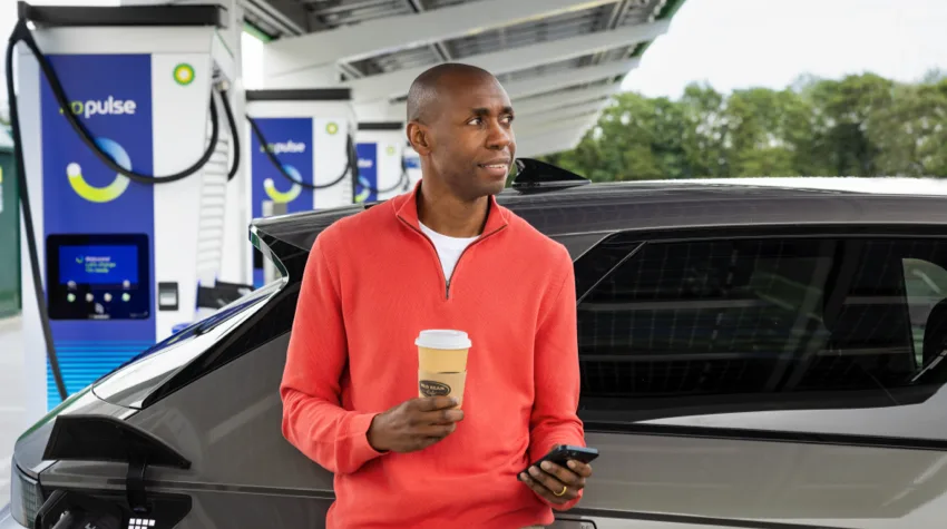 A man is standing with his back leaning on his dark silver Electric Vehicle. He is holding a wild bean coffee in one hand and has his mobile in the other hand. His Electric Vehicle is plugged in and charging, he is looking into the distance with a smile on his face.