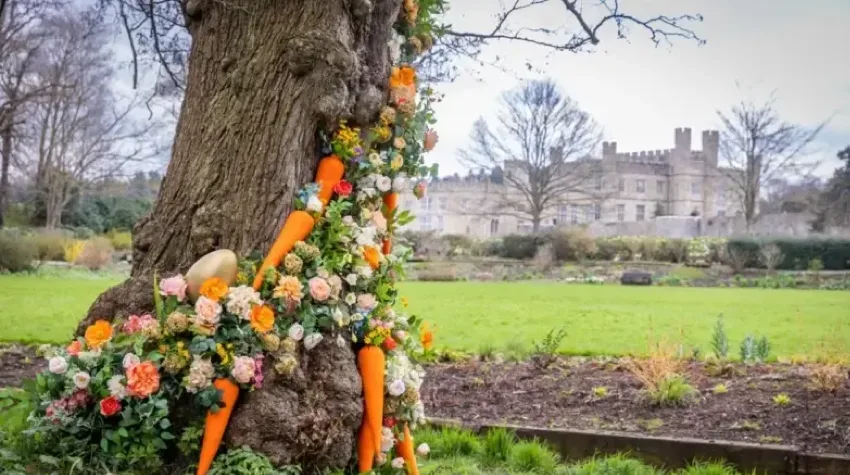 A large tree trunk decorated with vibrant spring flowers and orange carrots, set in the lush green grounds of the historic Leeds Castle in Kent.