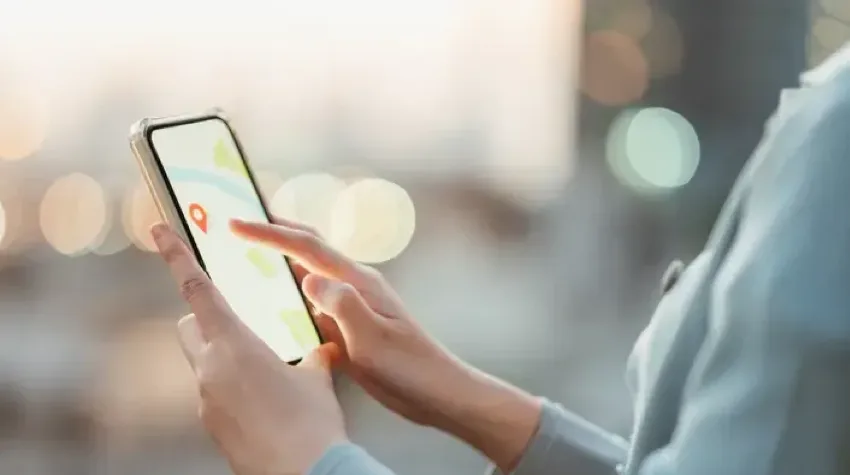 A person using a smartphone to navigate a map, with a red location pin visible on the screen and a blurred cityscape in the background.