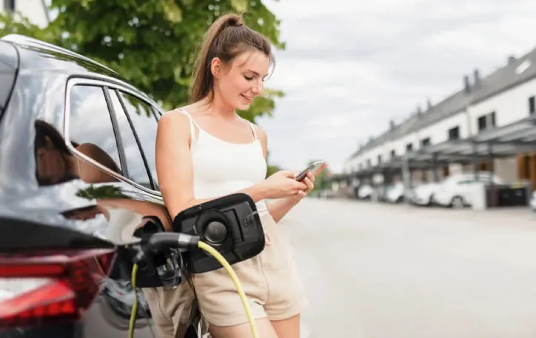 A woman leaning with her back against the side of a black Electric Vehicle looking at her phone, whilst the car is plugged in and charging.