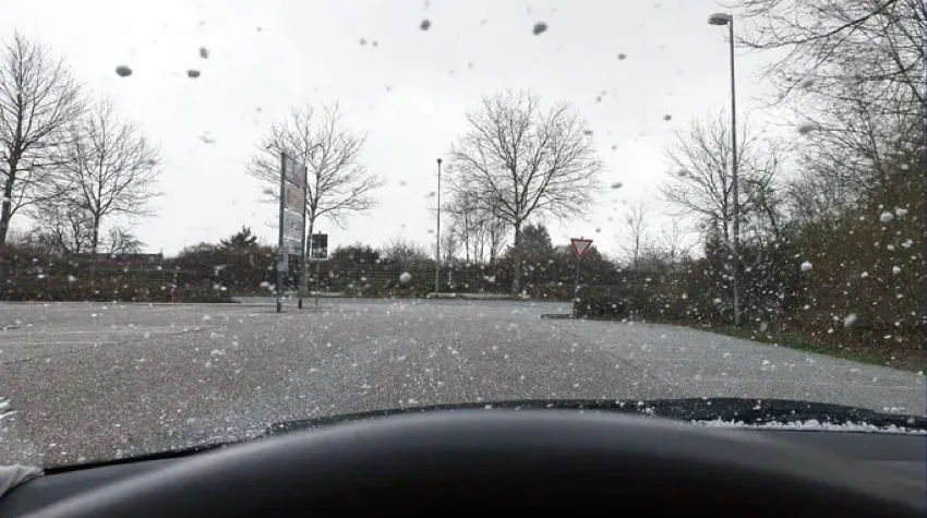 View of a hailstorm from inside a car, looking out through a windscreen covered in falling hailstones. An empty car park with bare trees and unlit street lamps and signs is visible under a grey, overcast sky.