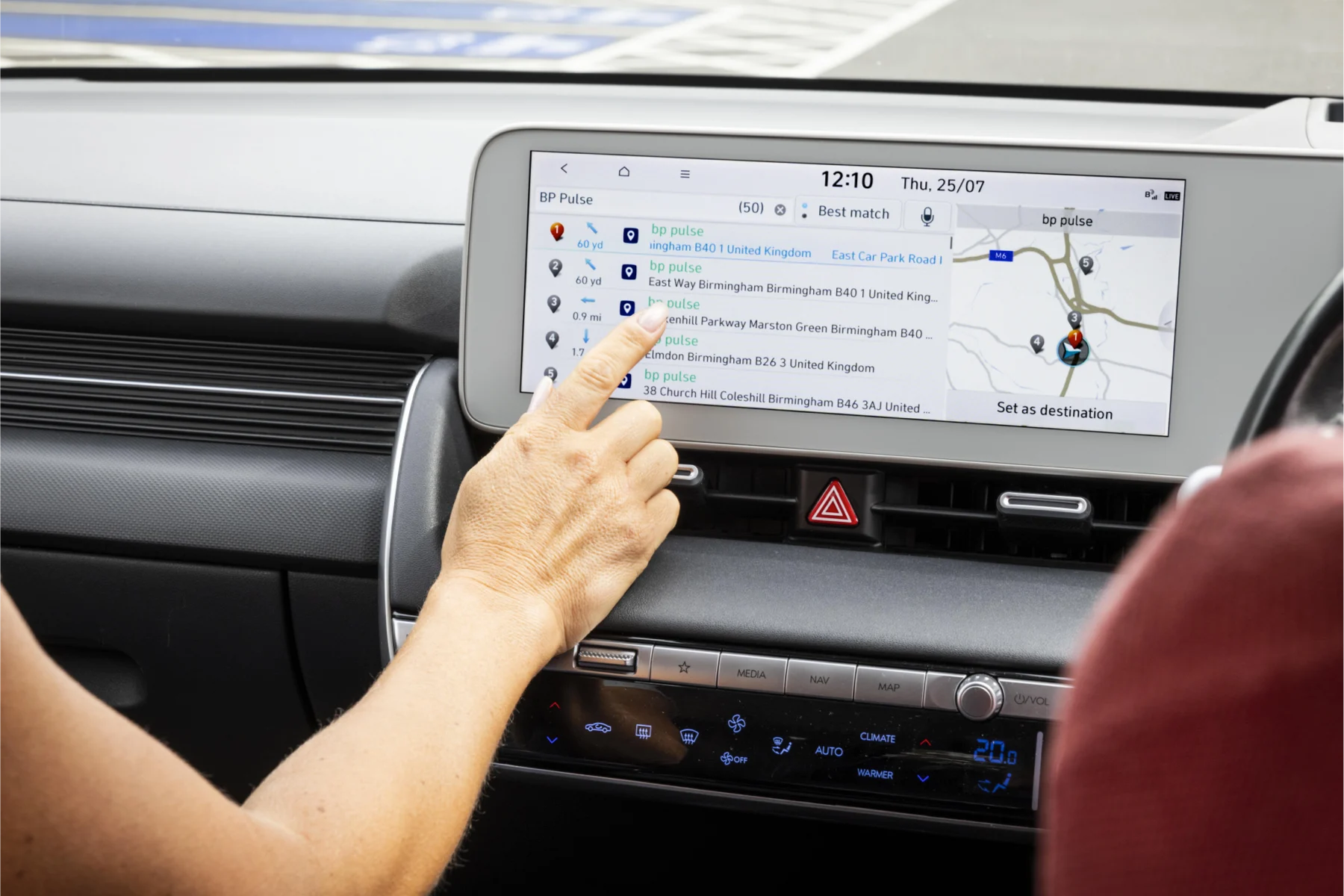 Close-up of a person's hand interacting with a car's touchscreen display, selecting a BP Pulse EV charging station from a list of nearby locations, with a map and route guidance shown on the right side of the screen.