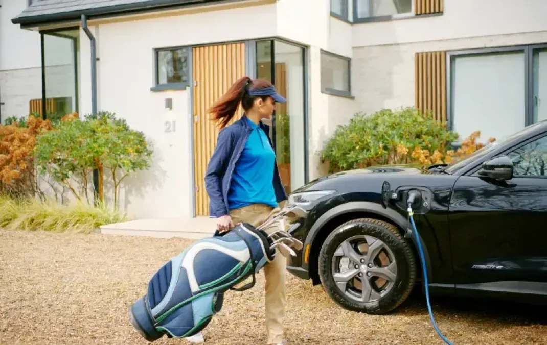 A woman is walking with her golf clubs towards her Electric Vehicle. The car is plugged in and charging in the driveway of a modern house.