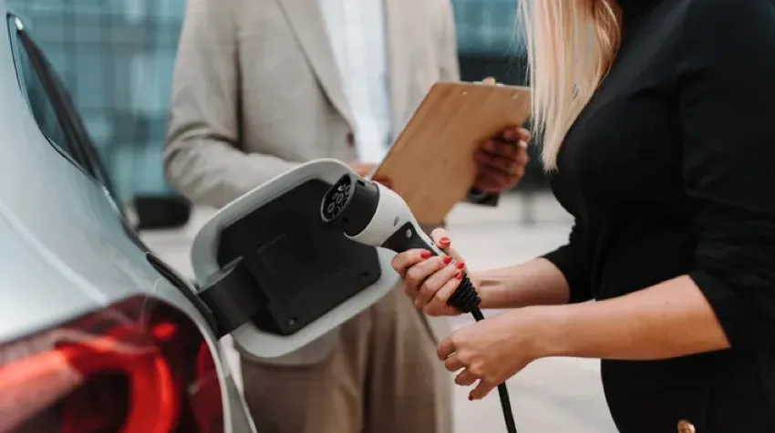 A man with a clipboard in his hand and a woman with an electric car charger in her hand, stand next to an electric car.
