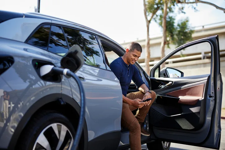 A person sits in the driver's seat of a parked electric vehicle, holding a smartphone while the car charges at a station. The vehicle's door is open, and a charging cable is connected. Trees and an overpass appear in the background.