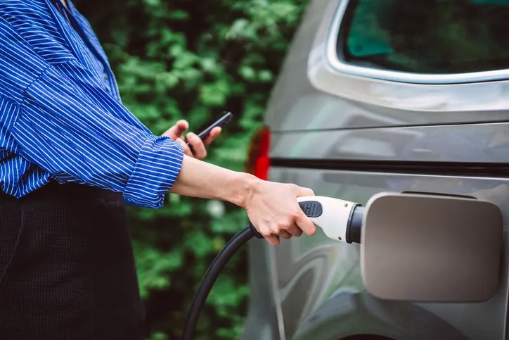A woman is connecting her grey EV to a charging socket.
