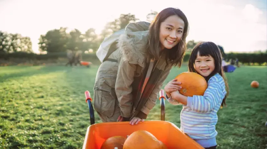 A lady and a little girl with a pumpkin posing for a picture in a park on bright day.