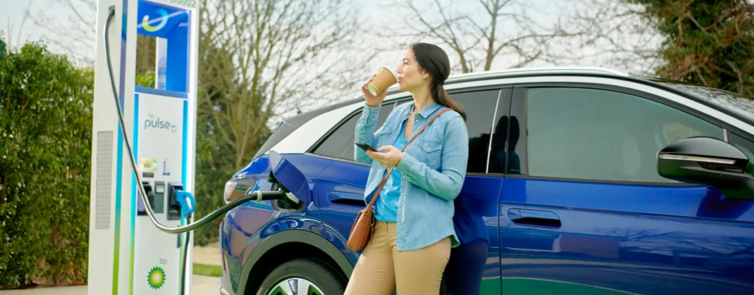A woman in a blue denim shirt sips coffee next to her blue EV, which is charging at a bp pulse charging point. 