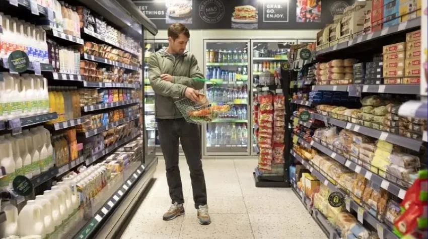 A man shopping in the aisle of M&S, he is placing an item into his shiopping basket.