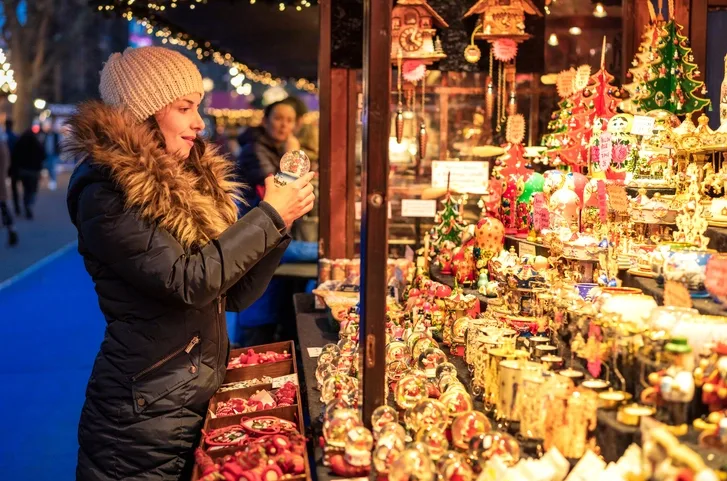 A lady wearing navy winter coat with fur colar and cream wooly hat is stood infront of a Christmas Market stall selling Christmas ornaments. The lady is looking at a snow globe that she is holding in her hands.
