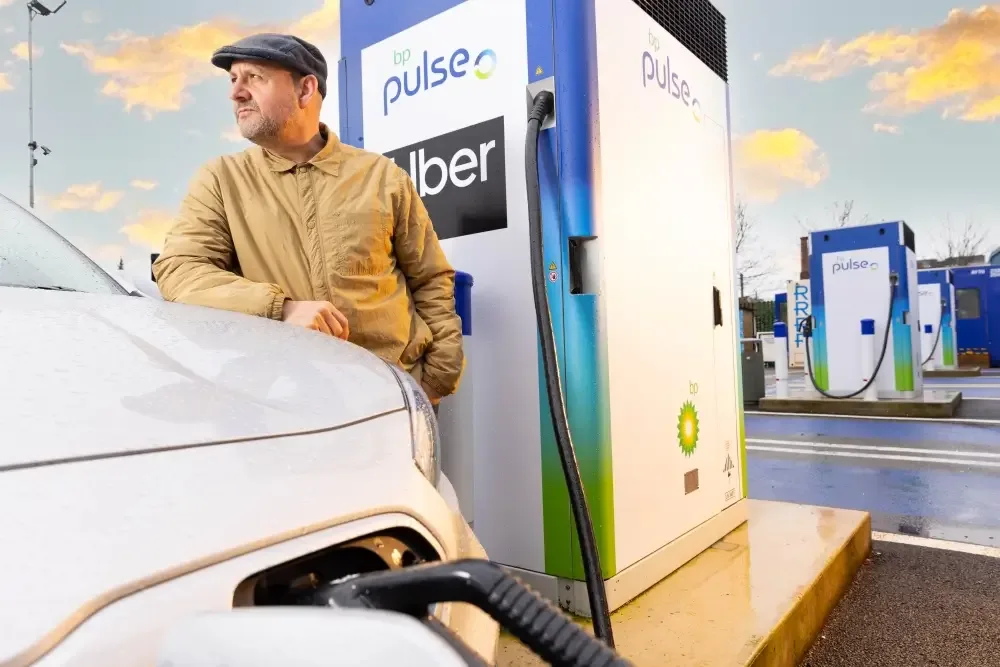 A photo of an Uber driver standing by his car wearing a flat cap. The car is next to a bp pulse charger.
