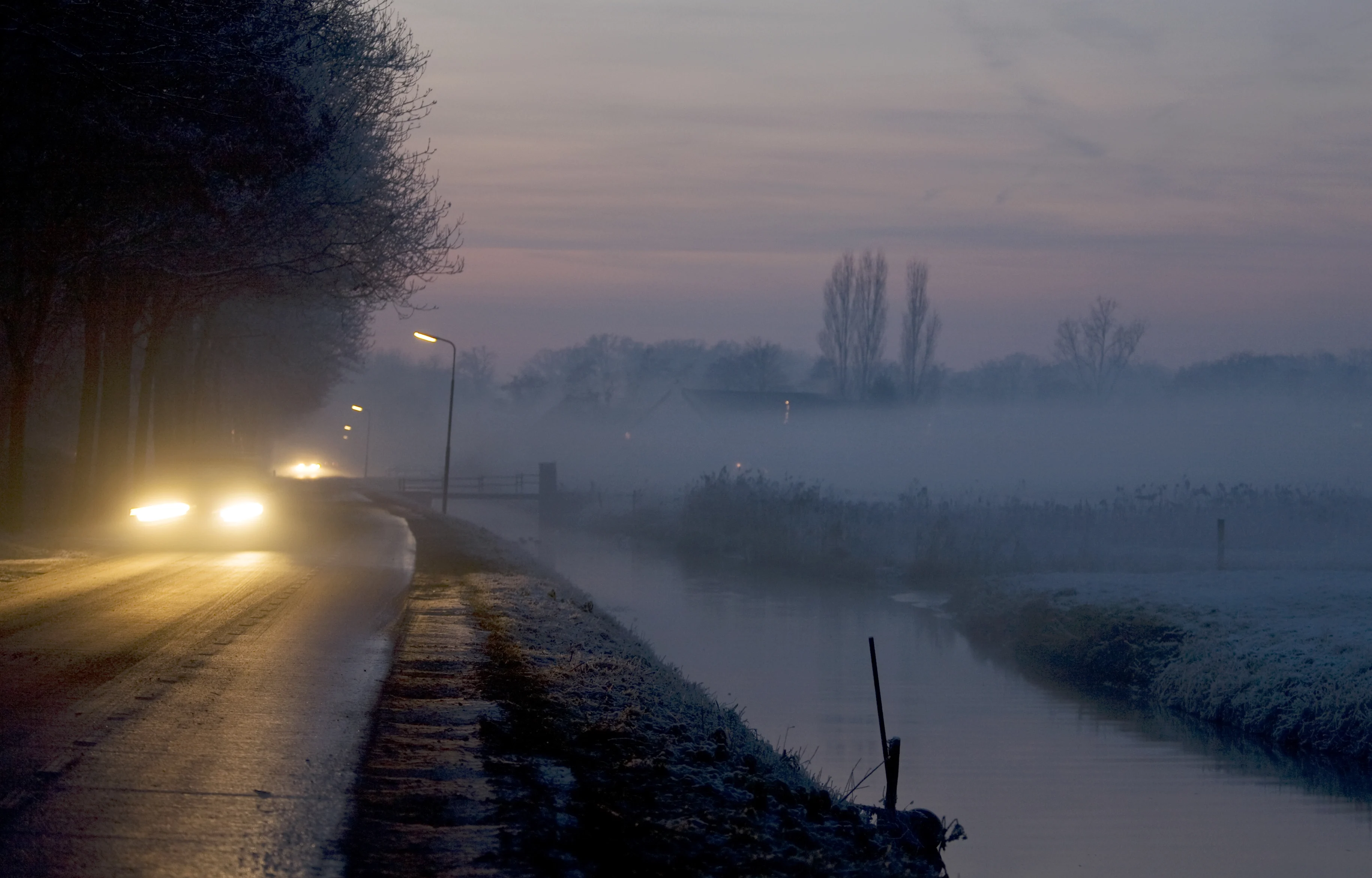 A car drives in the fog on a road next to a river.