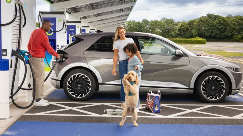 A father is charging his electric car at a bp pulse charging station while his wife, daughter, and dog are playing right next to the car.