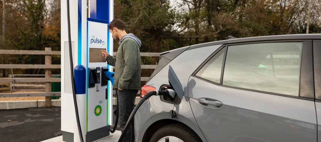 A man is tapping some information on a digital screen of a bp pulse charging station while an electric charger is plugged to his car.