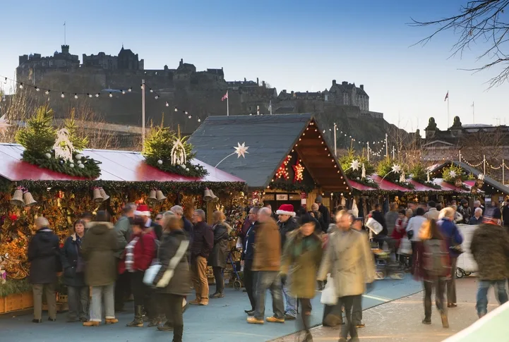 Edinburgh Christmas Market in the daytime with Edinburgh castle in the background. Lots of people dressed in winter clothing are walking around the market and appear blurred.