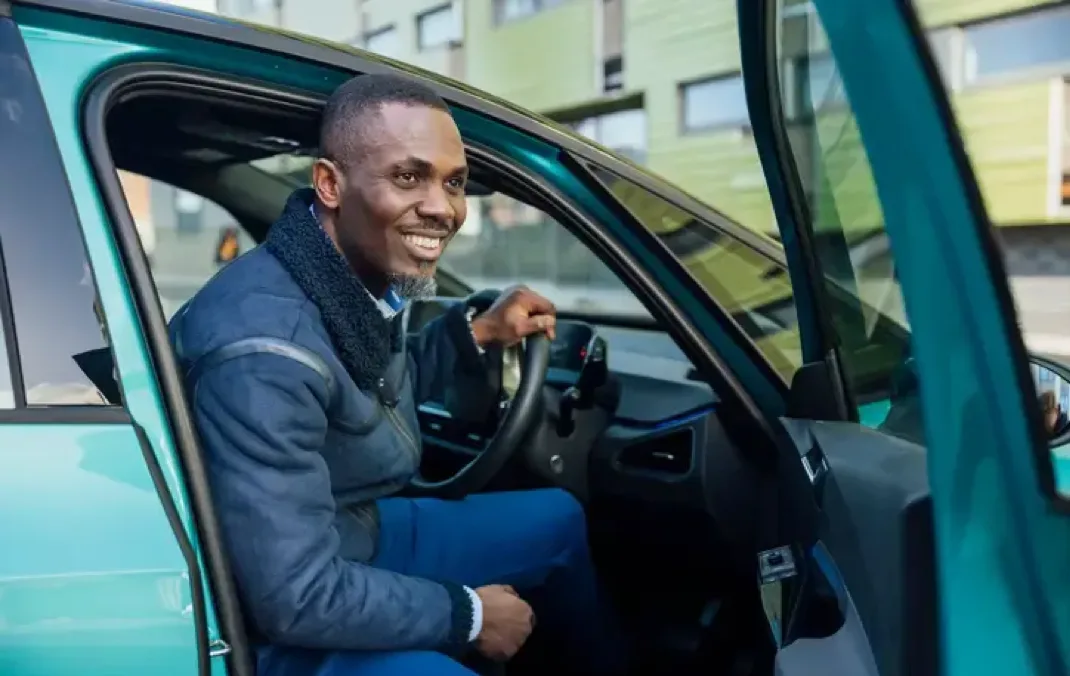 A smiling man, about to step out of a green car. The door is already open, and one of his hands is on the steering wheel. 