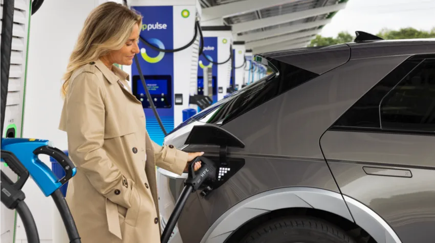 A woman in brown trench coat charging her grey electric car at a bp pulse charging station.