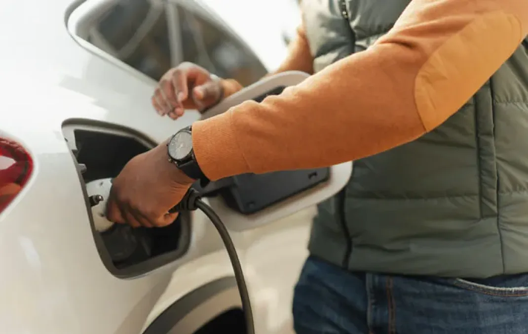 A man is plugging a charger into his white Electric Vehicle. His orange sleeve with a black wrist watch is visible.
