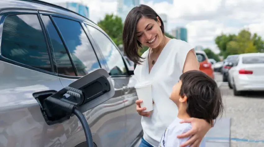 A mother and son standing next to their grey Electric Vehicle whilst it is charging. The mother is holding a coffee in one hand and has her other hand around her son on his shoulder. They are smiling affectionately at each other.