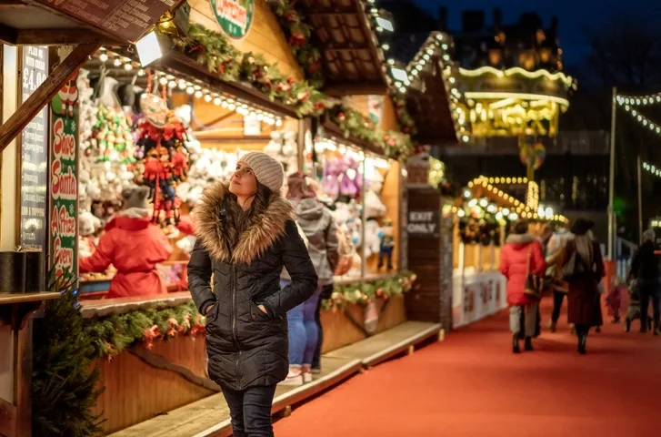 A woman in a navy winter coat with a fur collar and cream wooly hat with her hands in her pockets, is stood reading a menu on the walls of one of the Winchester Christmas Market huts. 