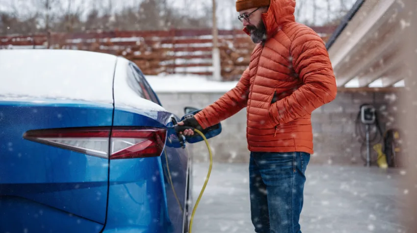 A man in an orange jacket charging his blue electric car in snowfall.