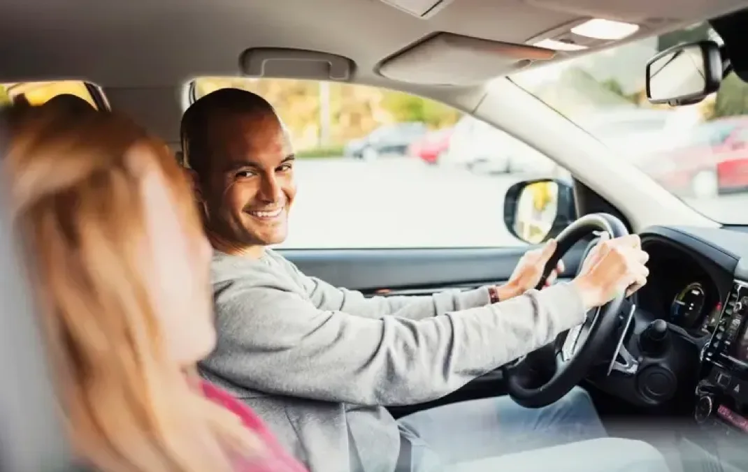 A man sitting in the driver seat of an EV. He has both hands on the steering wheel and is looking and smiling at the woman sat next to him in the passenger seat. The woman is looking at him.