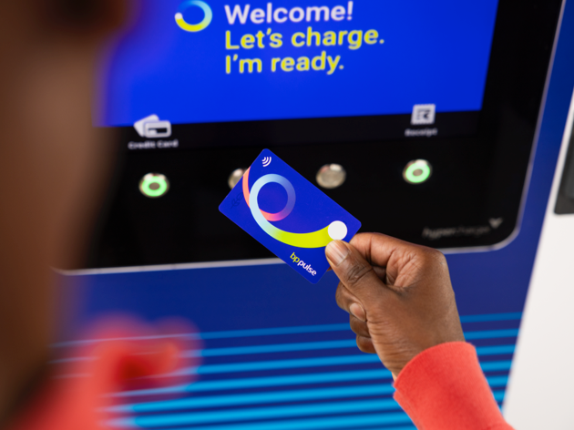 A close up of a man holding a bp pulse charge card between thumb and forefinger close to a bp pulse EV charger screen. The screen reads 'Welcome! Let's charge. I'm ready.'