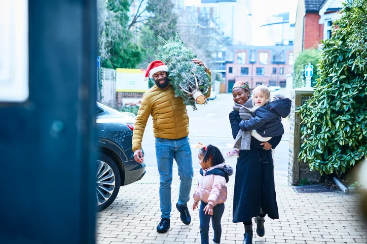 Cheerful mid adult man wearing Santa hat carrying Christmas tree on shoulder, arriving home with family