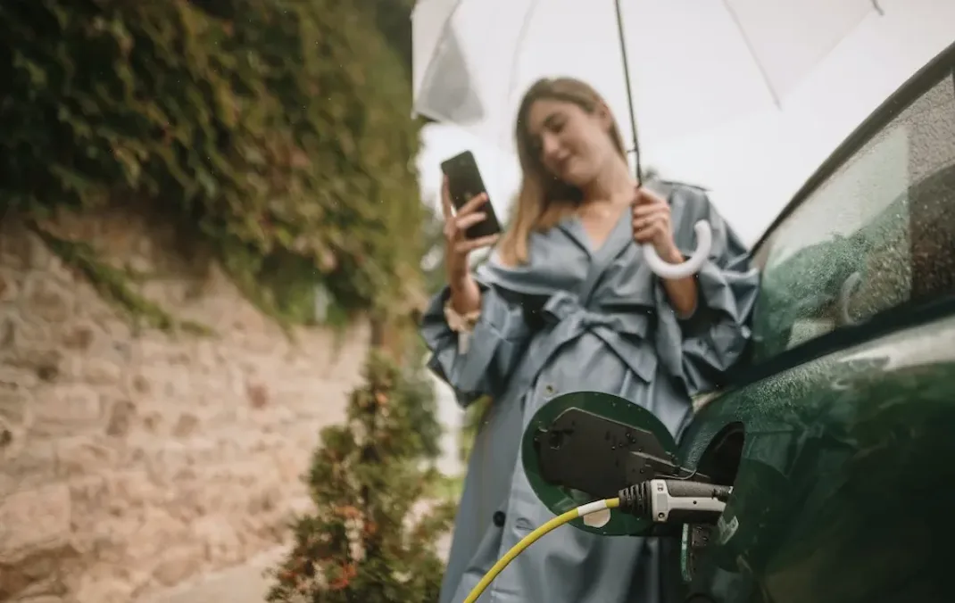 A woman leaning on her car, with a phone in one hand and an umbrella in another, while her car is being charged.