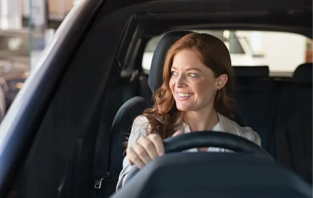 A woman sitting in an Electric Vehicle, she is sitting in the driving seat and has one hand on the steering wheel. She is smiling and looking out of the window.