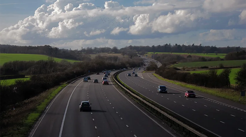 Cars driving on a multi-lane highway surrounded by green fields and trees under a cloudy sky.