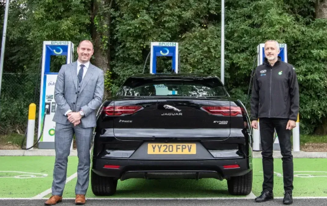 Member of Scottish Parliament Neil Gray and bp pulse EV Operations Lead Rob Chadwick stand next to a black Jaguar I-PACE in front of bp pulse chargers at Harthill services.