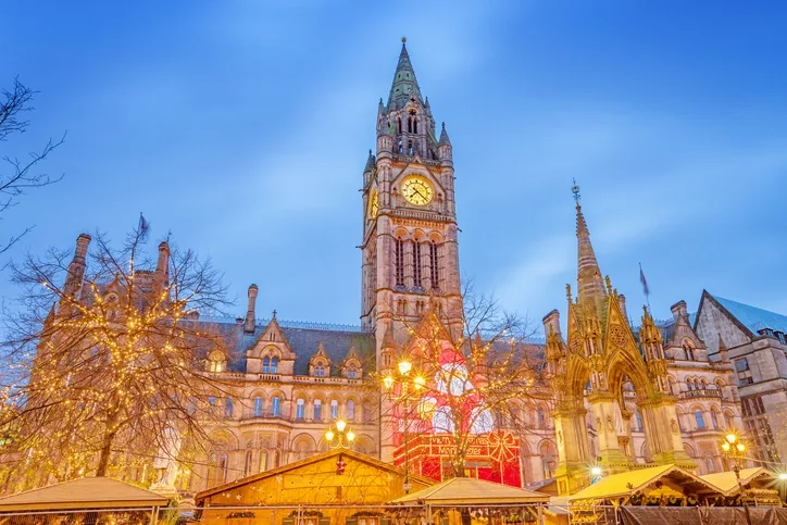Manchester Christmas Market in the early evening. A large lit up santa can be seen sat on a lit up present in front of the clock tower, with the tops of market huts in the forefront of the image. Two trees are lit with fairy lights giving off a warm glow.