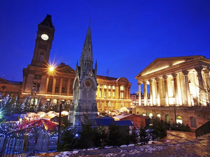 An evening photo of Birmingham Christmas market taken from the top of steps leading down to the market. The sky is a dark blue with the buildings lit up with soft glowing lights.