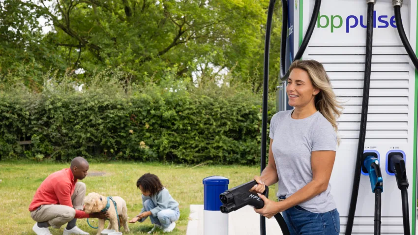 A woman stands next to a bp pulse electric vehicle charger, holding the charging connector in her hand. In the background, her family is crouched down on the grass, smiling and petting their dog.