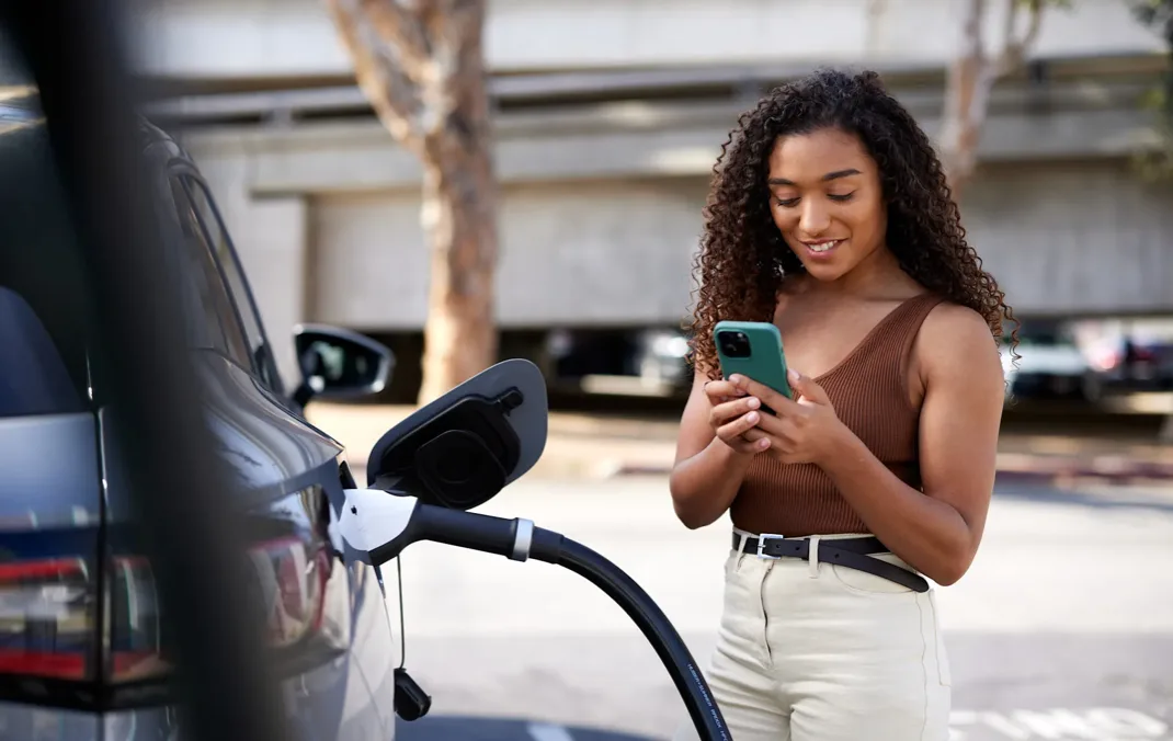 A woman is smiling and looking at her phone whilst her silver Electrical Vehicle is plugged in and charging. It is a bright sunny day.