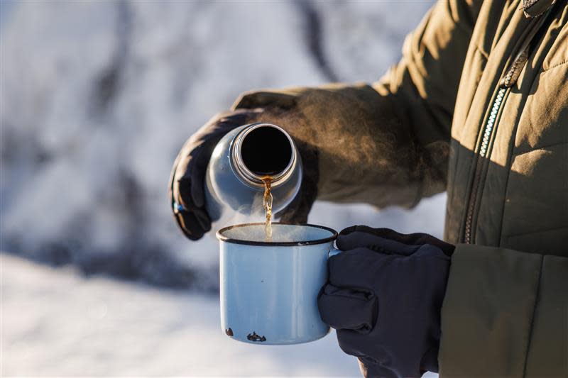 A person wearing gloves and a jacket pouring a hot beverage from a thermos into a blue enamel mug outdoors in a snowy environment.