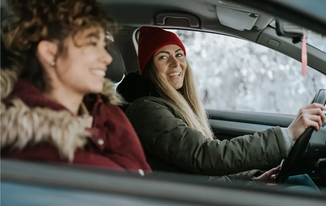 Two women dressed in winter clothing are sitting in the front seats of an Electric Vehicle. The driver is wearing a red woolly hat and has both hands on the steering wheel. She is smiling and looking across to the other woman, who is also smiling.