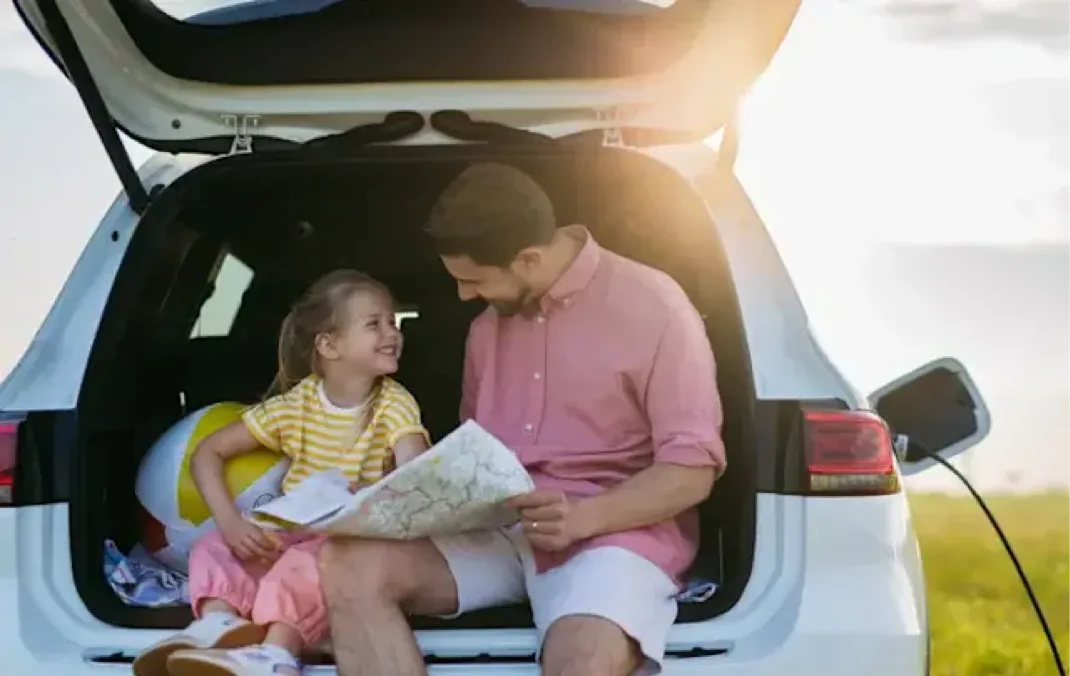 A father and daughter sit in the open boot of a charging car, smiling and looking at a map together.