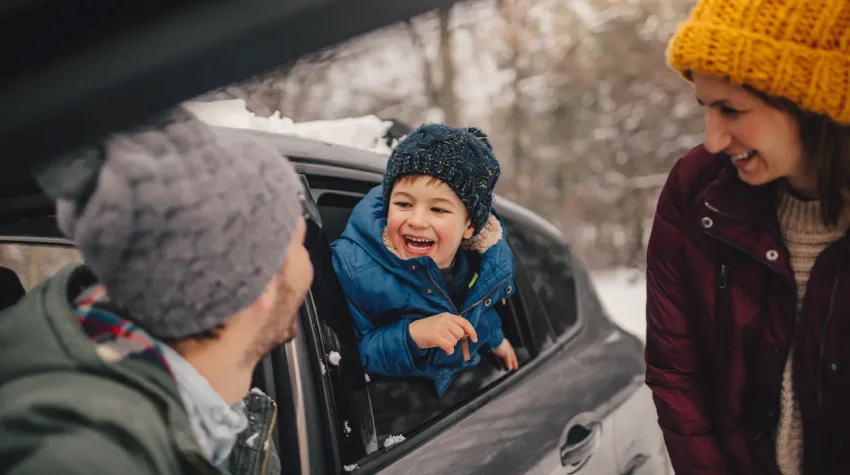 A smiling child is with his family, hanging out the side of their parked EV, in the snow. 