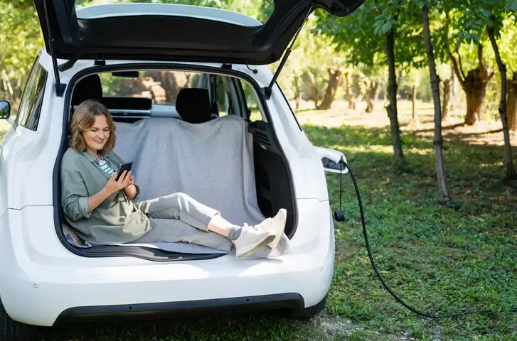 A photo of a young woman sitting in the trunk of her car looking at her mobile phone. Her car is being charged.