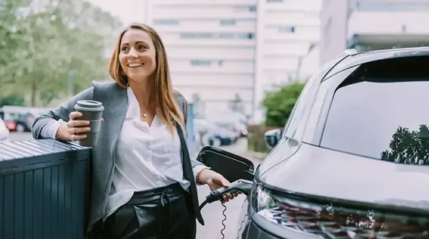 A lady in formal clothes is smiling, while she holds a coffee mug in her right hand and EV charger in her left hand to charge her car.
