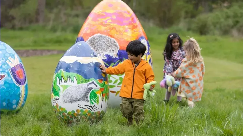 Three children exploring large, painted Easter eggs in RHS Garden Bridgewater, Manchester. The eggs feature colourful nature-themed artwork, including animals and landscapes.